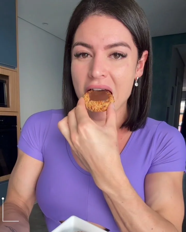 Vallery comendo uma bolachinha fit com chocolate, de blusa roxa, em vídeo de receita